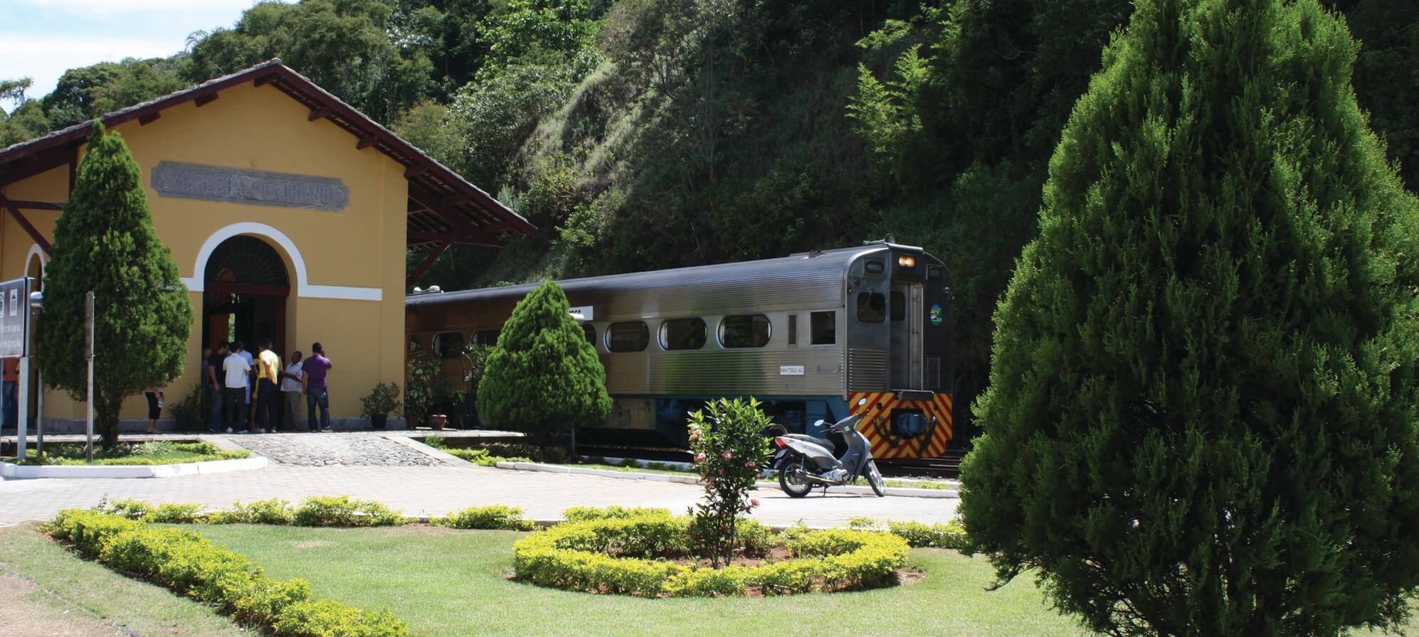 Serra Verde Express train arriving at a scenic station near Curitiba, surrounded by gardens and forested hills. 