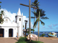 Historic white church by the sea in Praia do Forte, Brazil, with palm trees and a clear blue sky. 