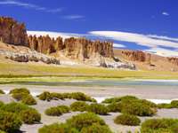 Dramatic rocks spires rise above green shrubs and a salt flat in Chile's high altitude Altiplano region. 
