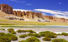 Dramatic rocks spires rise above green shrubs and a salt flat in Chile's high altitude Altiplano region. 