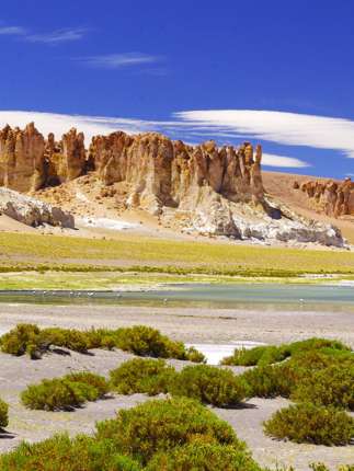 Dramatic rocks spires rise above green shrubs and a salt flat in Chile's high altitude Altiplano region. 