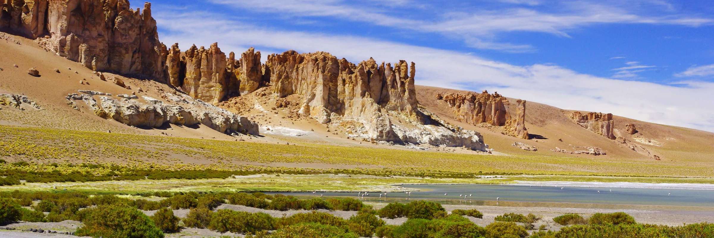 Dramatic red rock formations rise above a high-altitude lagoon and wetlands in Chile's Atacama Altiplano under a vivid blue sky. 