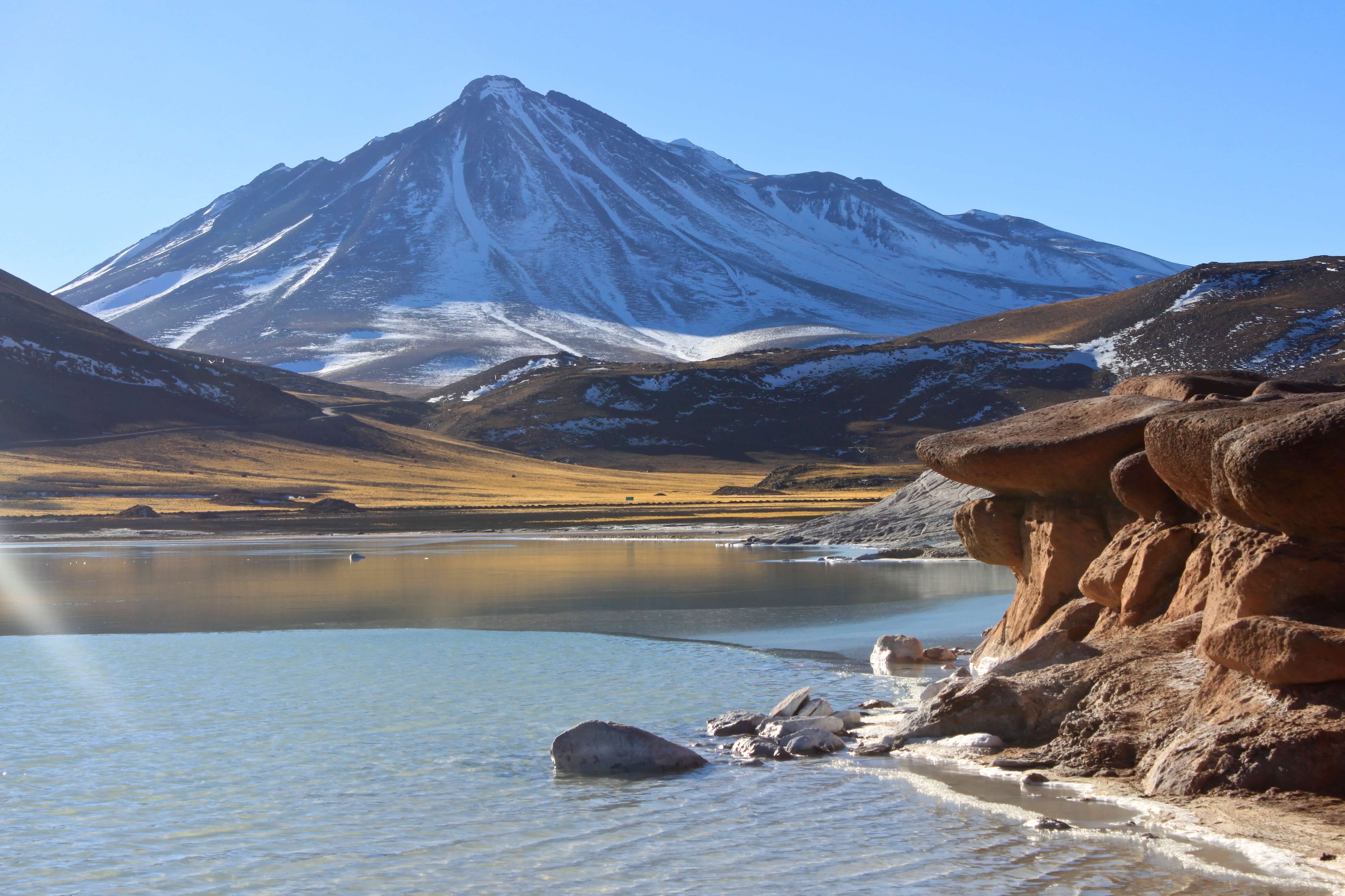 Snow-capped Licancabur volcano rises above a serene Atacama lagoon with sculpted rock formations in the foreground. 