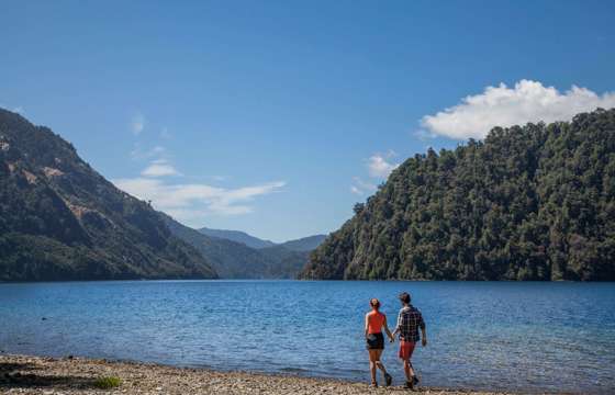 A couple walks along the shore of a clear lake surrounded by forested hills in Chile's Huilo Huilo Biological Reserve. 