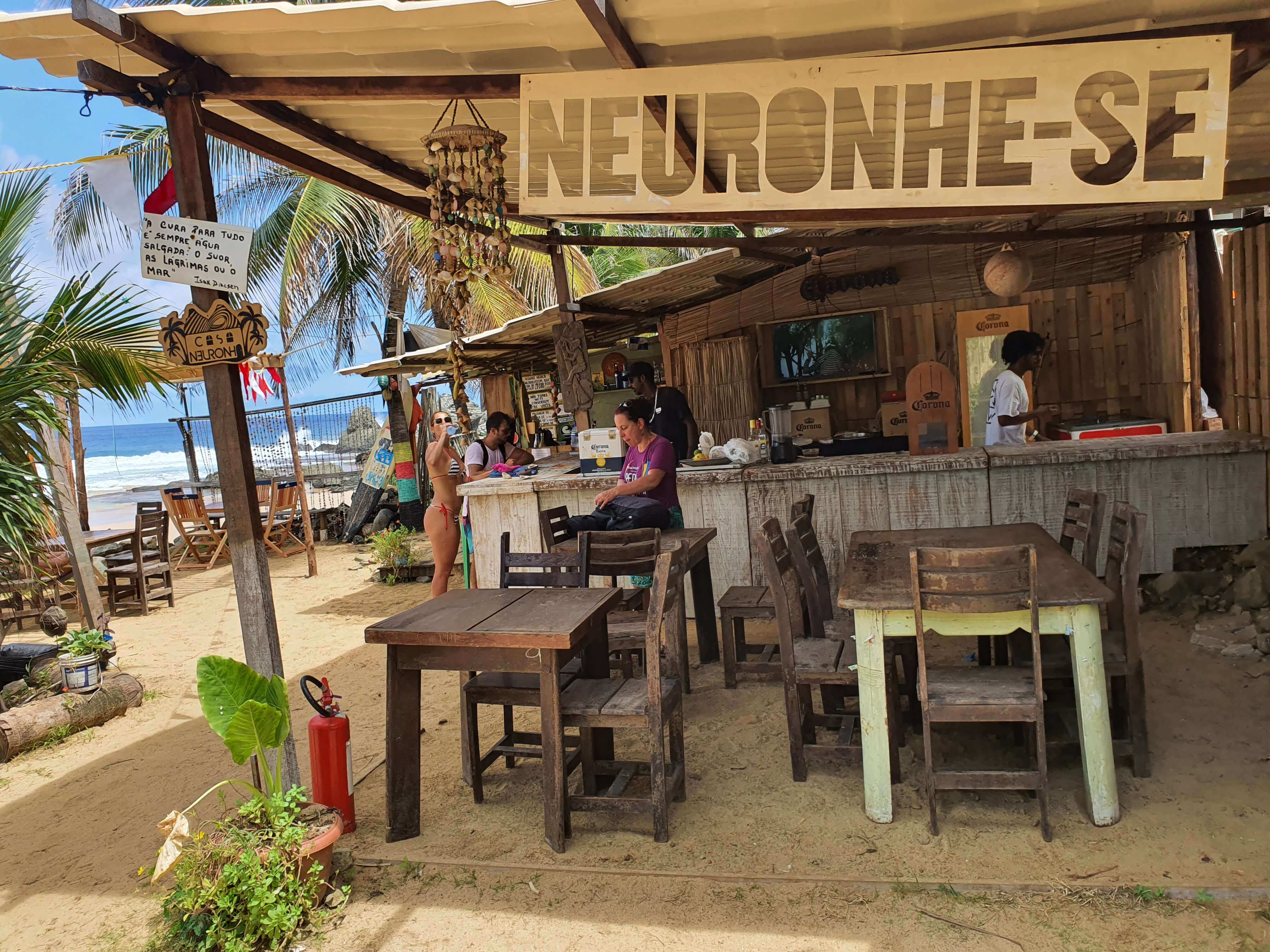 Rustic beachfront bar with wooden furniture and palm trees in Fernando de Noronha, Brazil, featuring a Neuronhe-se sign. 