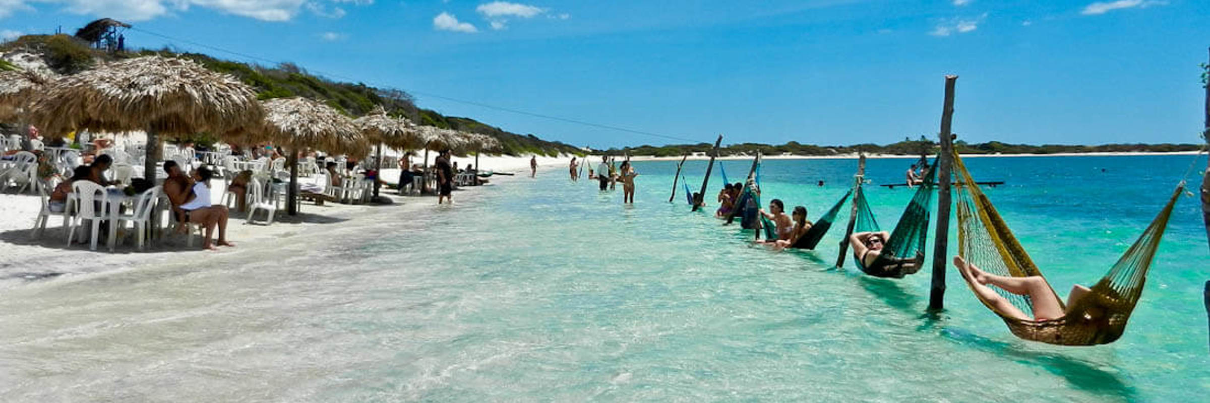 Tourists relaxing in hammocks over clear waters at a lagoon in Jericoacoara, surrounded by dunes and blue skies. 