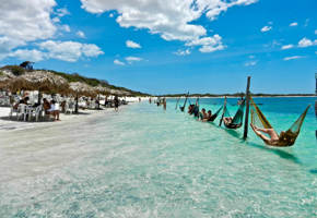 Tourists relaxing in hammocks over clear waters at a lagoon in Jericoacoara, surrounded by dunes and blue skies. 