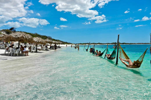 Tourists relaxing in hammocks over clear waters at a lagoon in Jericoacoara, surrounded by dunes and blue skies. 