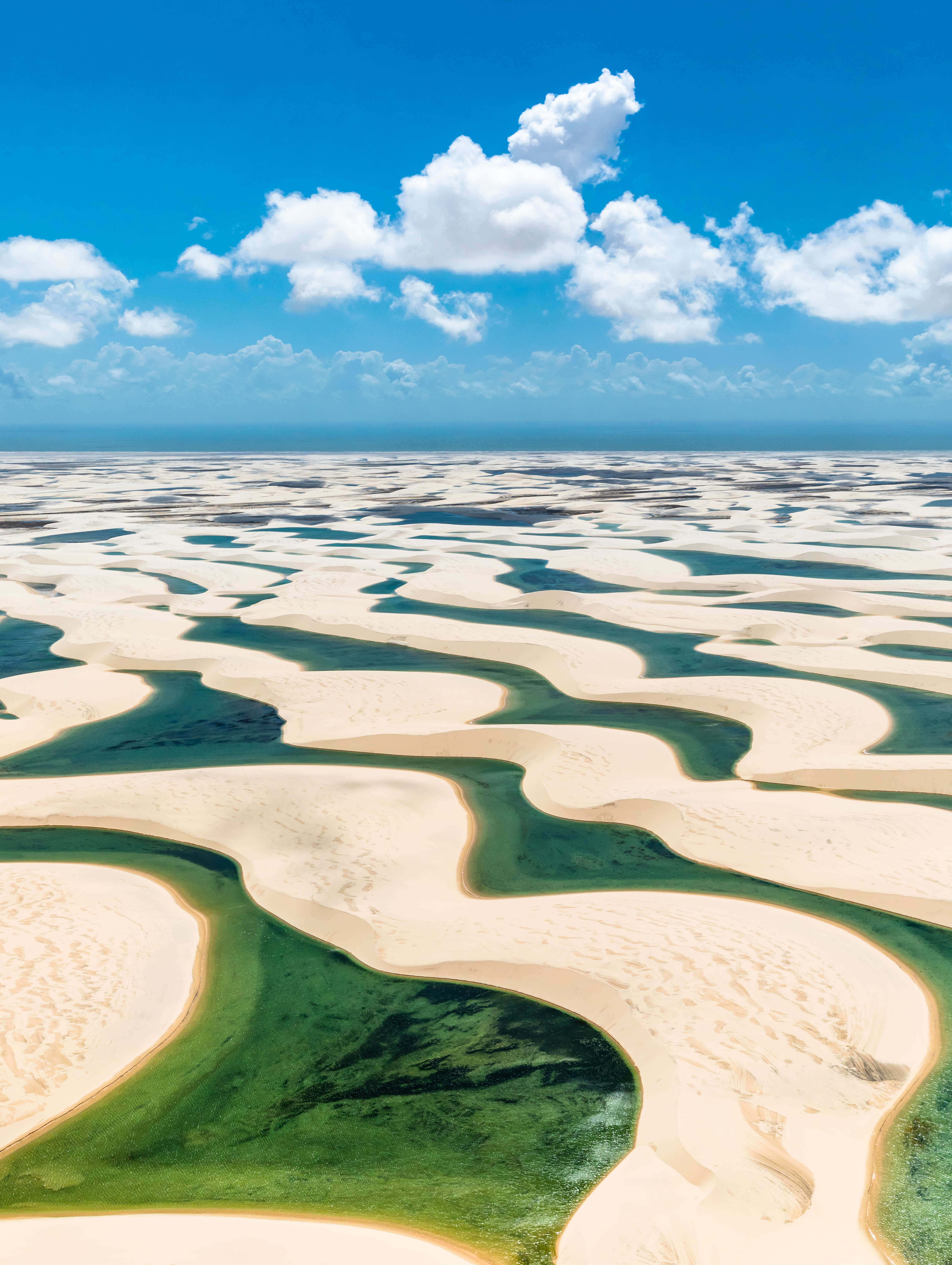 Aerial view of Lencois Maranhenses' white sand dunes and emerald lagoons beneath a vibrant summer sky. 