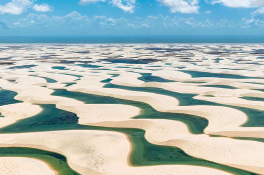 Aerial view of Lencois Maranhenses' white sand dunes and emerald lagoons beneath a vibrant summer sky. 