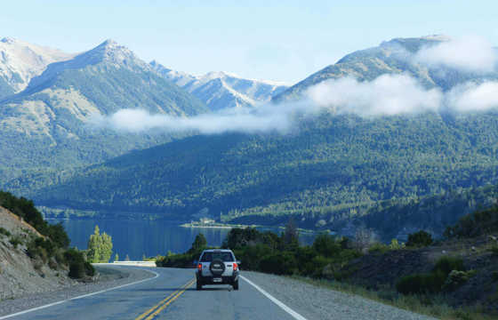 Car drives through winding road in Chile's Lake Region, flanked by lush mountains and low-lying mist under a clear sky. 