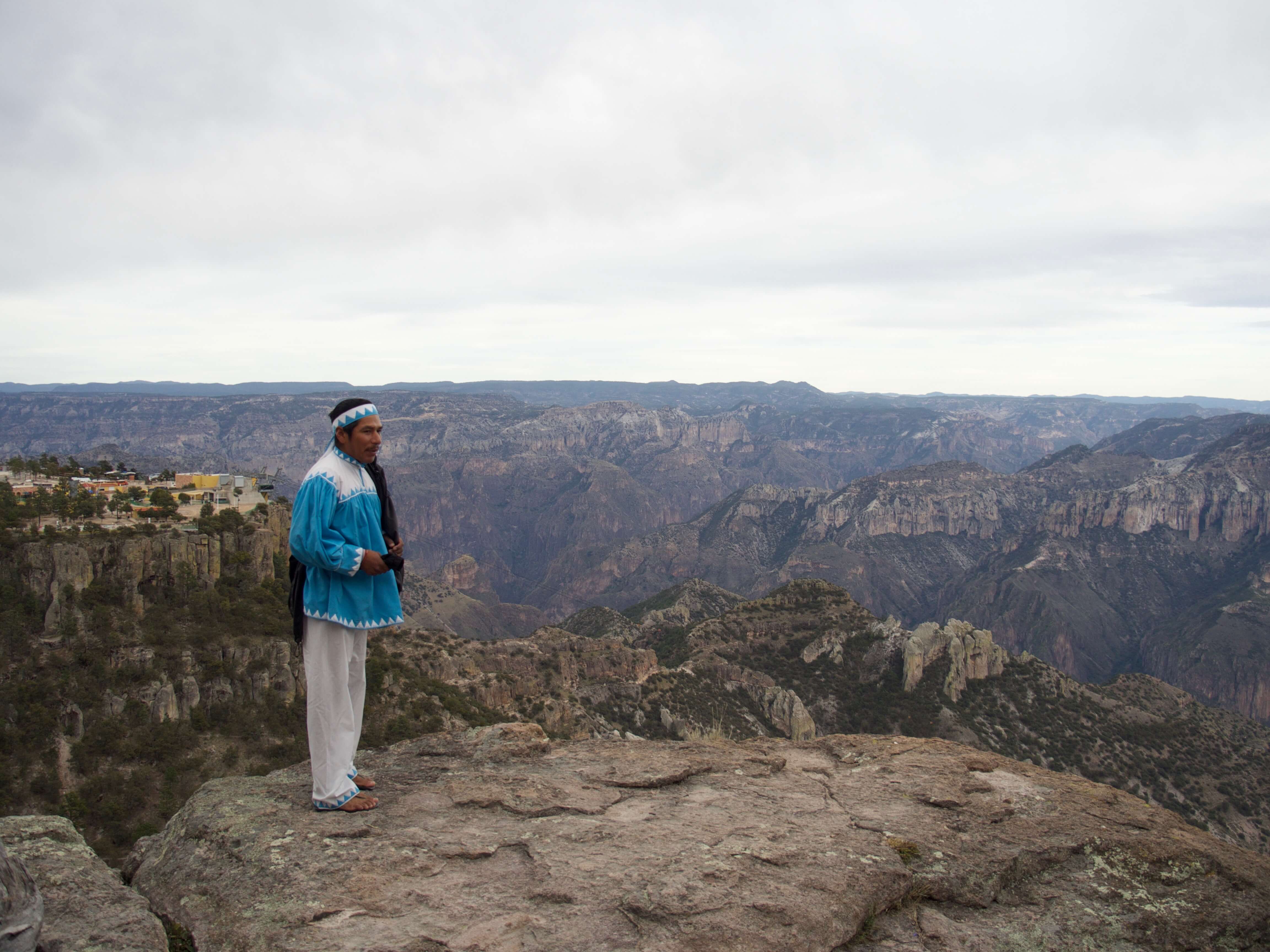 Tarahumara man in traditional clothing stands on a cliff edge overlooking the dramatic Copper Canyon in northern Mexico. 
