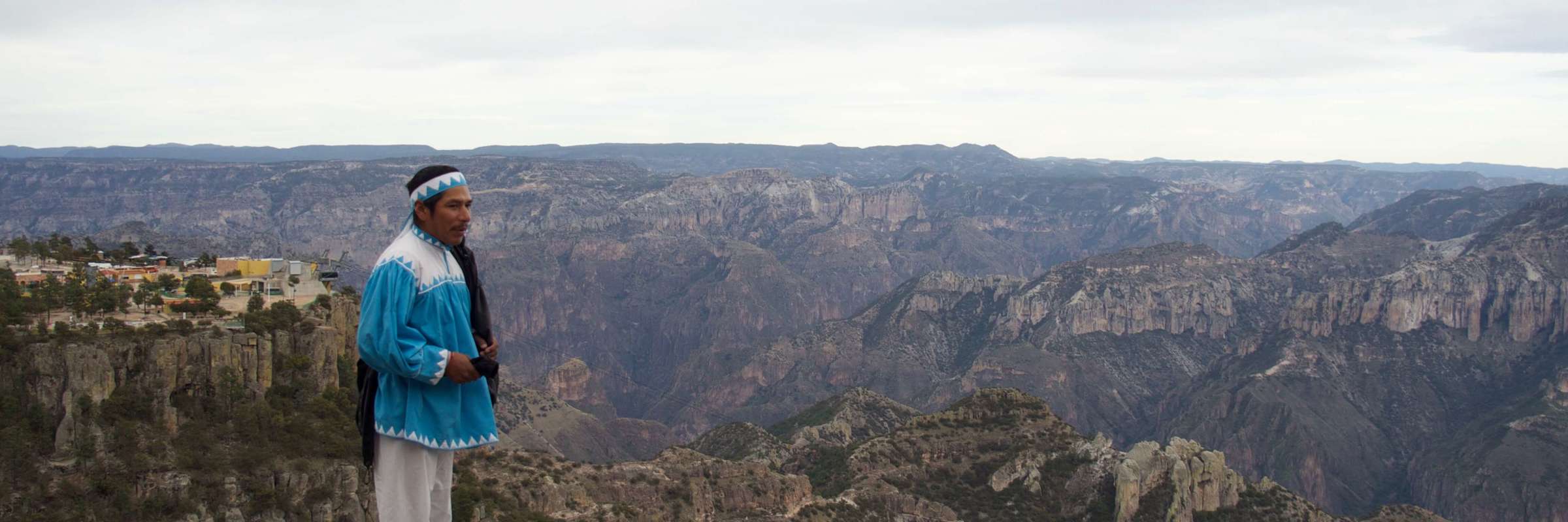 Tarahumara man in traditional clothing stands on a cliff edge overlooking the dramatic Copper Canyon in northern Mexico. 