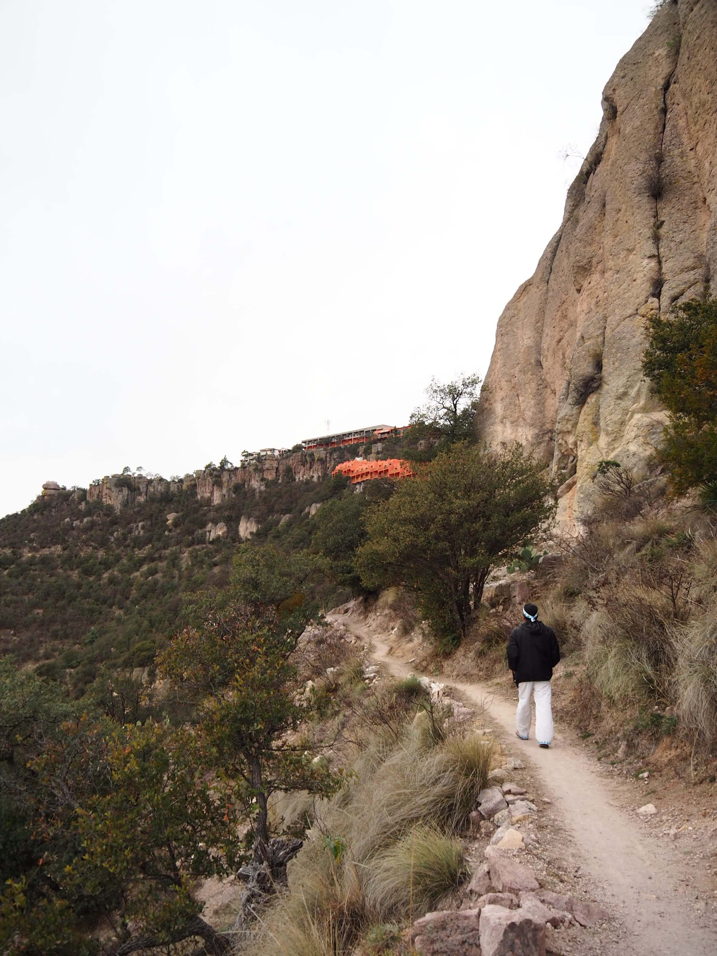 Tarahumara man walking a mountain trail in Copper Canyon with the El Chepe train visible on the cliffs in the background. 