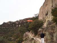 Tarahumara man walking a mountain trail in Copper Canyon with the El Chepe train visible on the cliffs in the background. 