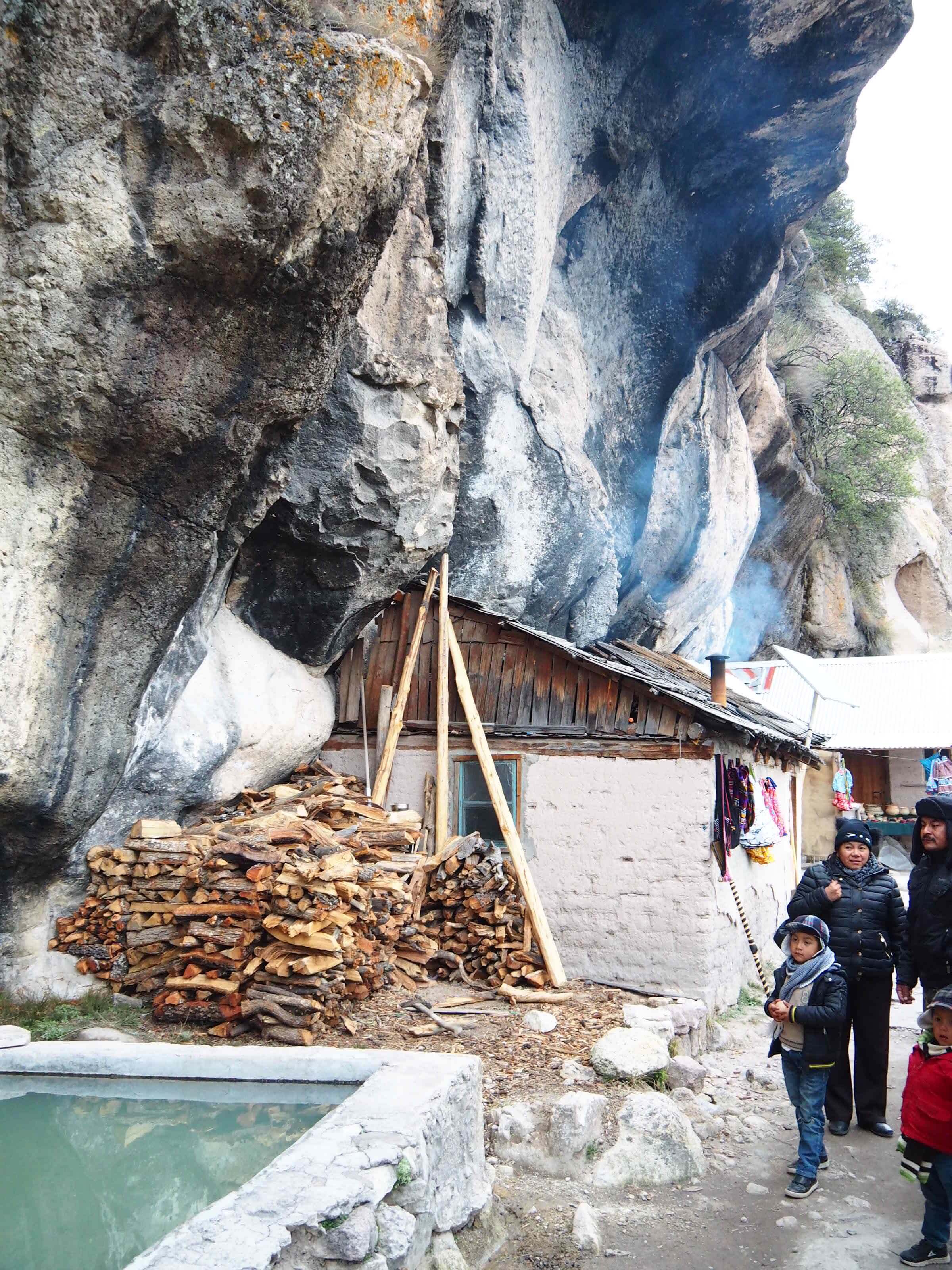 Local Tarahumara family near a rustic wooden hut and hot springs pool at Recowata, surrounded by cliffs in Copper Canyon. 