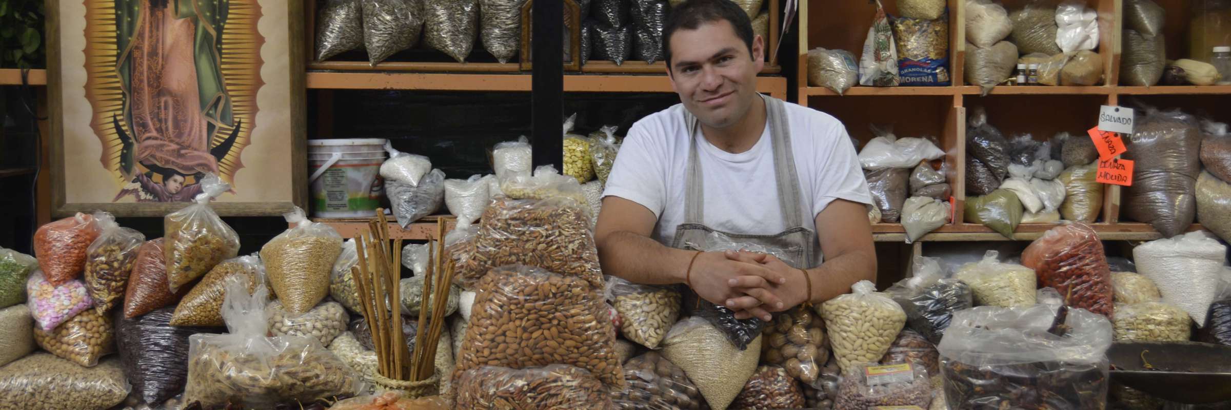 Friendly vendor in San Miguel de Allende sits among sacks of grains and spices, under the gaze of the Virgin of Guadalupe. 