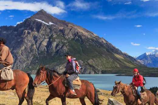 A group of horseback riders follows a scenic trail near a lake, with rugged Patagonian mountains rising in the background in southern Chile. 