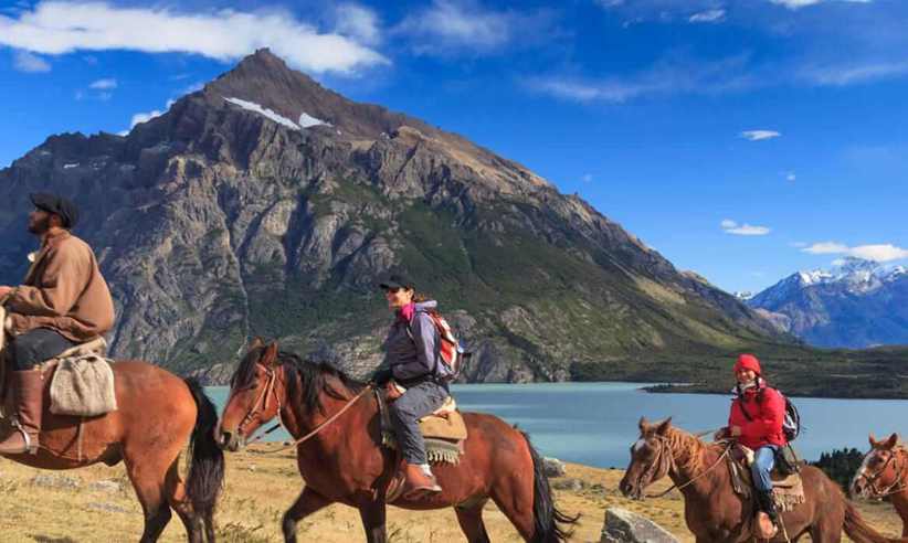 A group of horseback riders follows a scenic trail near a lake, with rugged Patagonian mountains rising in the background in southern Chile. 