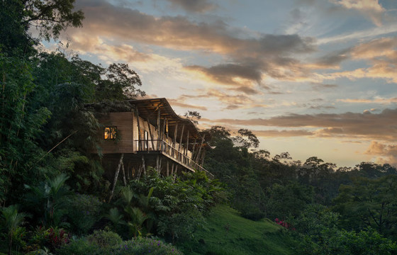 A luxury hillside villa at Origins Lodge overlooks lush forest and distant peaks, bathed in golden sunset light. 