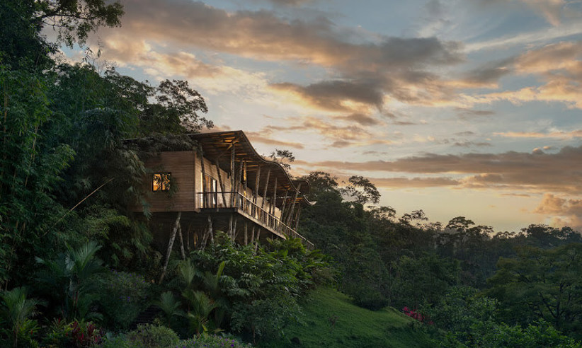A luxury hillside villa at Origins Lodge overlooks lush forest and distant peaks, bathed in golden sunset light. 