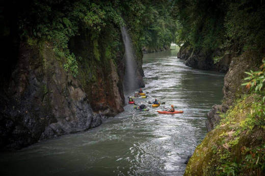 A group of Kayakers navigates the narrow canyon of the Pacuare River, surrounded by lush rainforest cliffs. 