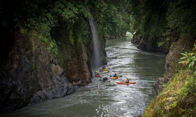 A group of Kayakers navigates the narrow canyon of the Pacuare River, surrounded by lush rainforest cliffs. 