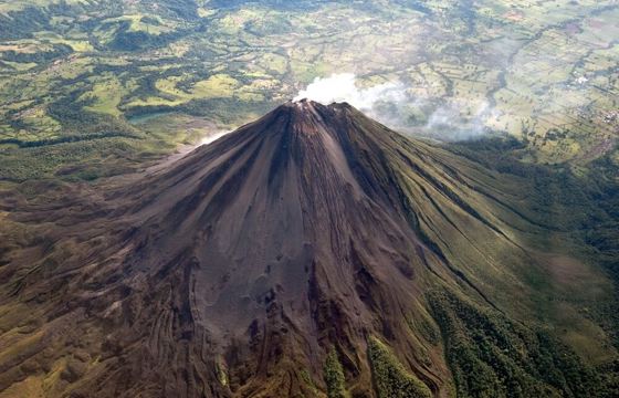 A dramatic aerial shot captures Arenal Volcano's perfect cone rising above rugged lava terrain. 