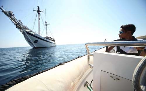 Man aboard a small boat approaches a traditional Indonesian sailing ship on calm waters during a luxury island-hopping cruise. 