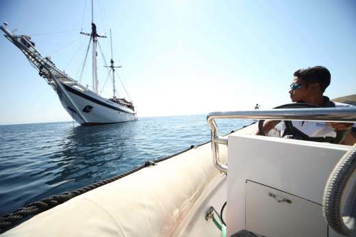 Man aboard a small boat approaches a traditional Indonesian sailing ship on calm waters during a luxury island-hopping cruise. 