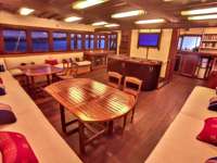 Warm wooden dining area on the Ombak Putik yacht, with long cushioned benches, vibrant pillows, and ambient lighting. 