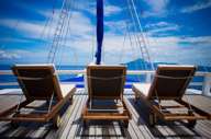 Three wooden loungers on the sun deck of a cruise ship, facing the open sea under a bright blue tropical sky. 