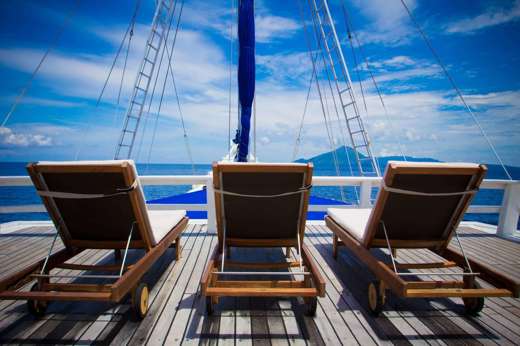 Three wooden loungers on the sun deck of a cruise ship, facing the open sea under a bright blue tropical sky. 