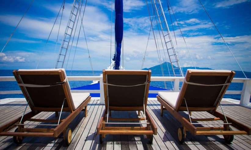 Three wooden loungers on the sun deck of a cruise ship, facing the open sea under a bright blue tropical sky. 