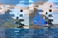 A traditional schooner with blue sails cruises past a towering volcano in Indonesia, capturing the essence of archipelago exploration. 