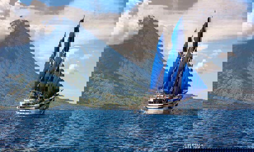 A traditional schooner with blue sails cruises past a towering volcano in Indonesia, capturing the essence of archipelago exploration. 