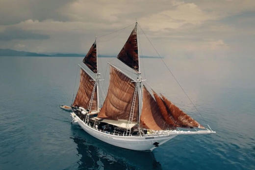 The Ombak Putik, a traditional Indonesian sailing ship, glides across calm blue waters under soft skies near the Komodo Islands. 