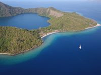 An aerial view of Satonda Island, Indonesia, showing a lush green coastline, crater lagoon, and deep blue ocean with a single white boat. 