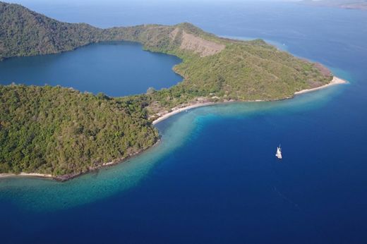 An aerial view of Satonda Island, Indonesia, showing a lush green coastline, crater lagoon, and deep blue ocean with a single white boat. 