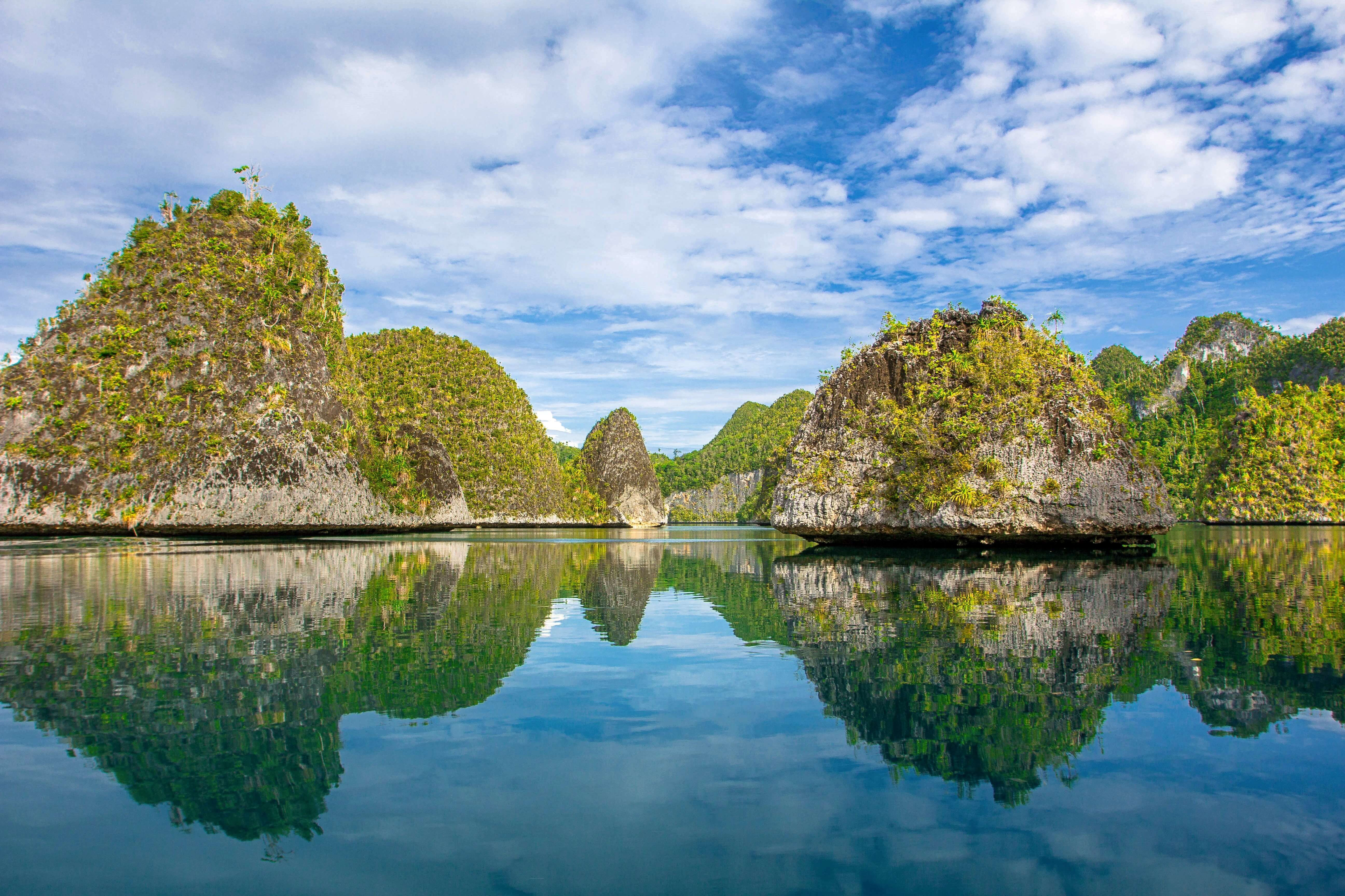 Crystal-clear waters reflect limestone islets and blue skies in Misool, Indonesia, creating a perfect mirror-like tropical seascape. 