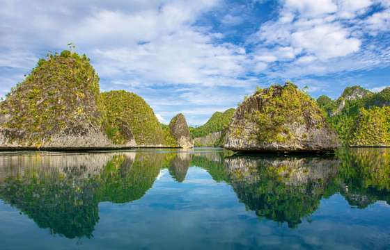 Crystal-clear waters reflect limestone islets and blue skies in Misool, Indonesia, creating a perfect mirror-like tropical seascape. 