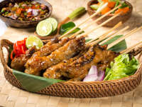 Traditional Indonesian chicken sate skewers served in a woven basket with fresh lime, vegetables, and spicy sambal on the side. 
