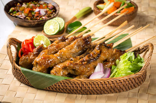 Traditional Indonesian chicken sate skewers served in a woven basket with fresh lime, vegetables, and spicy sambal on the side. 