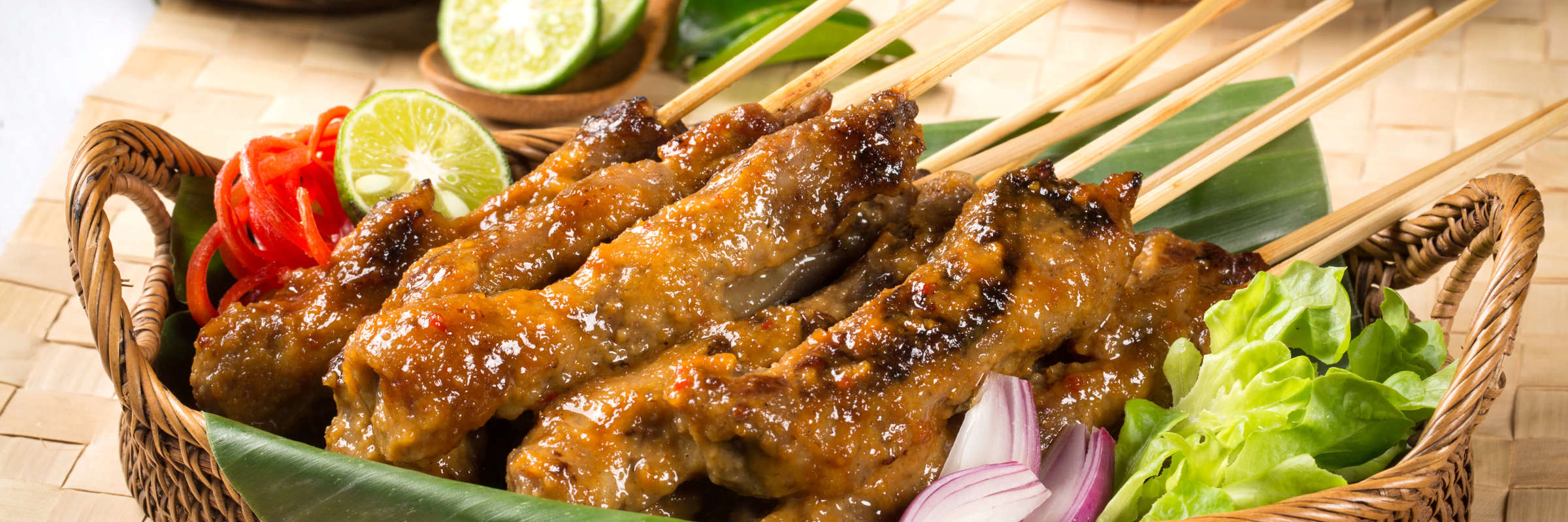 Traditional Indonesian chicken sate skewers served in a woven basket with fresh lime, vegetables, and spicy sambal on the side. 