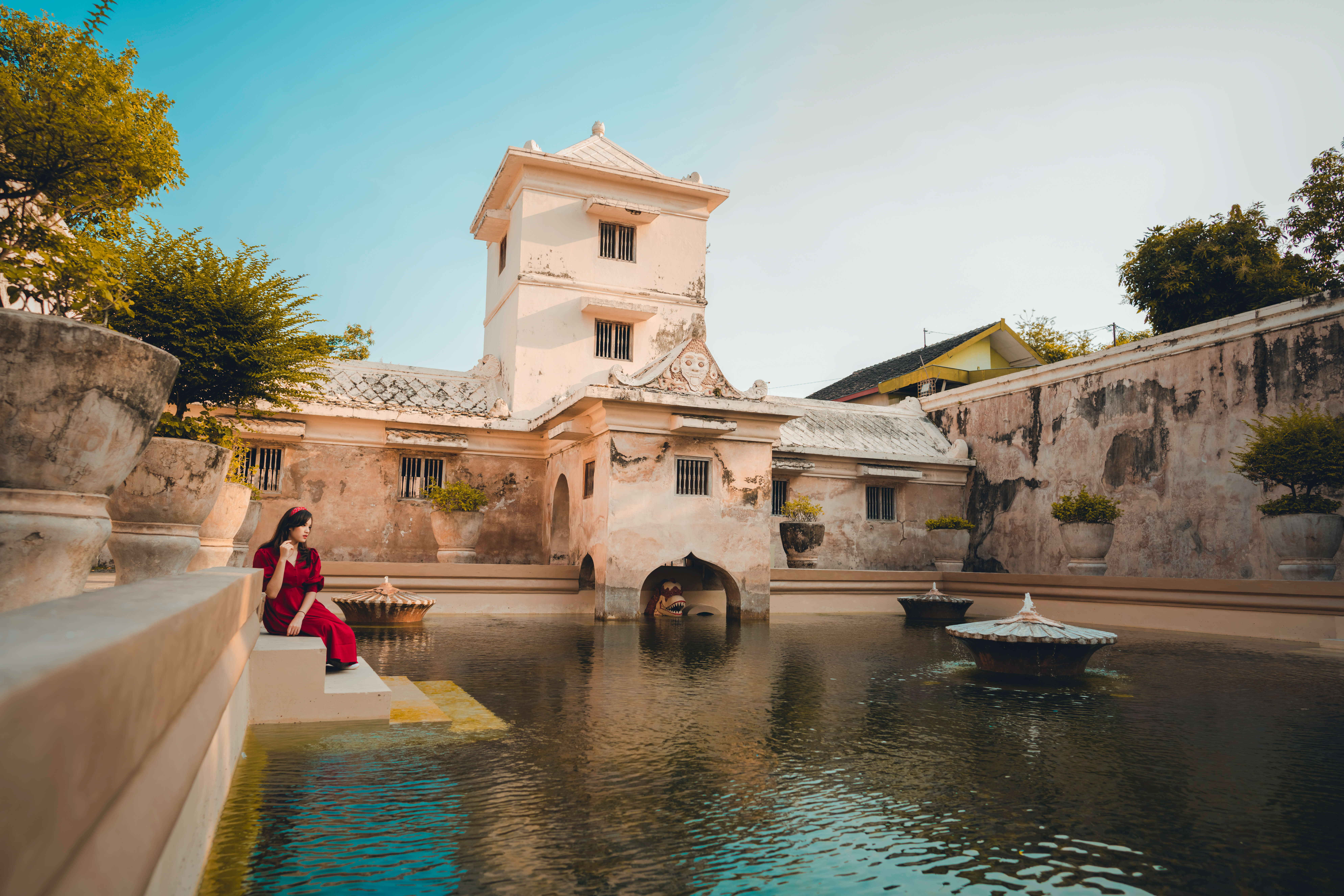 A woman in red sits by the bathing pool of Taman Sari Water Palace in Yogyakarta, Indonesia, framed by historic architecture. 