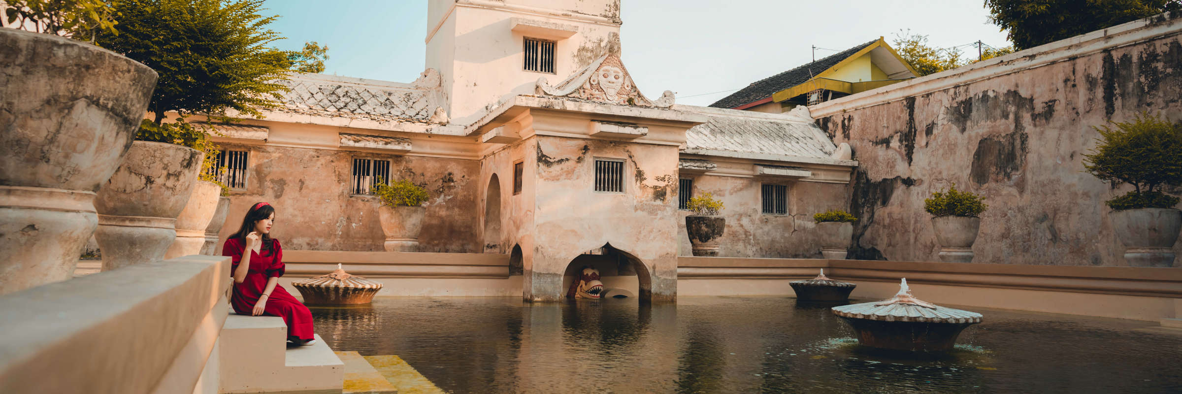 A woman in red sits by the bathing pool of Taman Sari Water Palace in Yogyakarta, Indonesia, framed by historic architecture. 