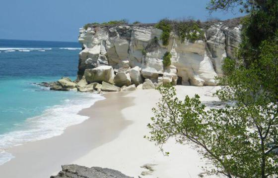 Turquoise waves meet the white sands of Sumba, backed by rugged limestone cliffs and lush greenery under a bright sky. 