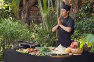 A Balinese chef stands with hands pressed in greeting beside a table of fresh ingredients during an outdoor cooking class. 