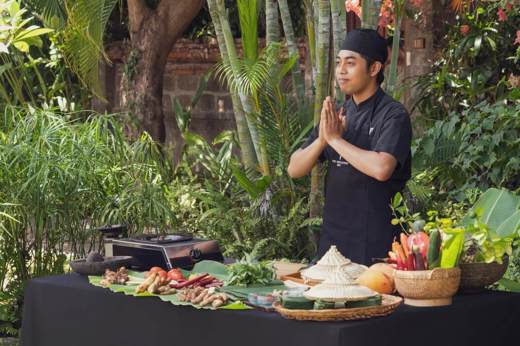 A Balinese chef stands with hands pressed in greeting beside a table of fresh ingredients during an outdoor cooking class. 