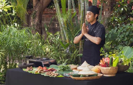 A Balinese chef stands with hands pressed in greeting beside a table of fresh ingredients during an outdoor cooking class. 
